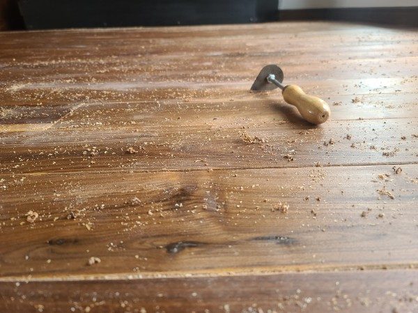 A hand scraper resting on a dark stained plank surrounded by fine shavings