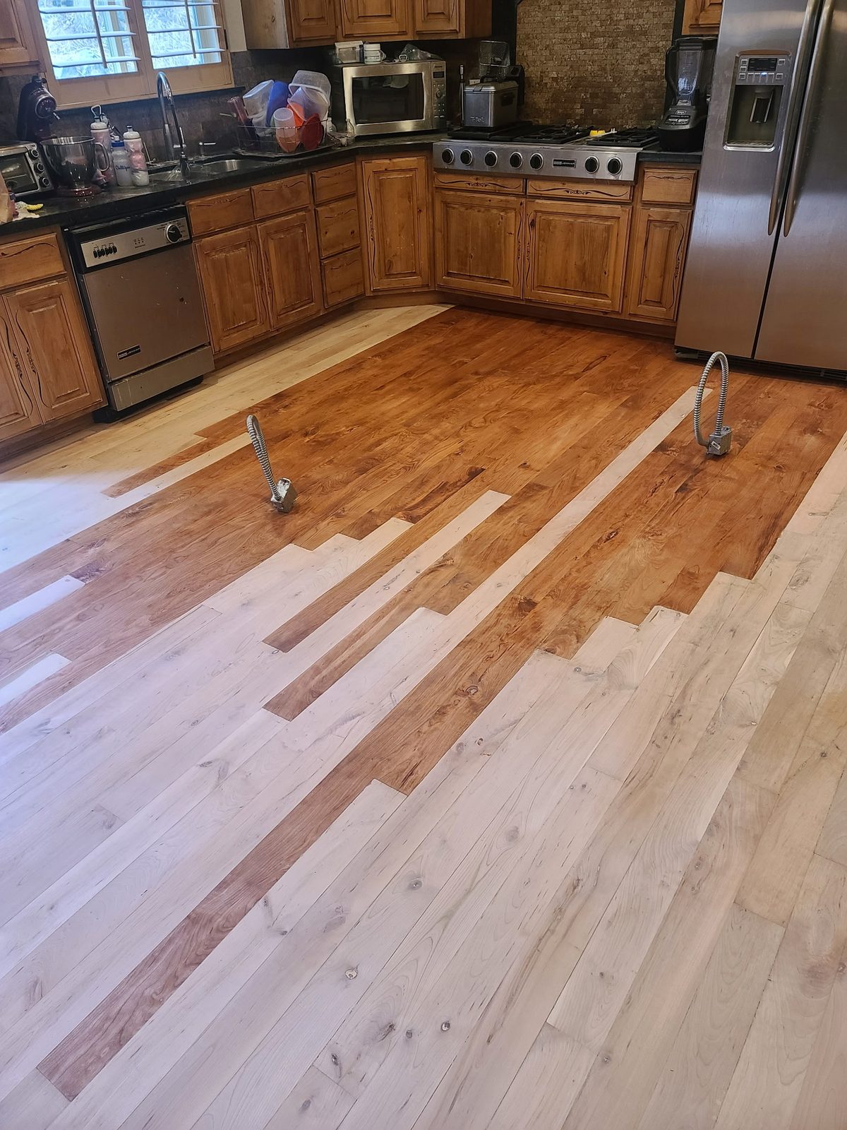 A kitchen floor mid-stain: the far half already carries a rich amber colour while the near boards are still bare, freshly sanded wood